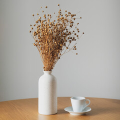 Cup of coffee and vase with flax dry grass on wooden table. White wall on background. Still life nordic, Scandinavian home decor. Cozy home living room interior.