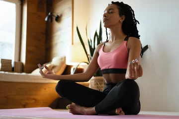 Schilderijen op glas Young black woman doing yoga at home in the lotus position. © opolja