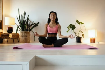 Fototapeten Young black woman doing yoga at home in the lotus position. © opolja