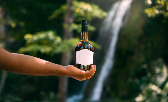 Men holds bottle with white label in front of waterfall in the summer.