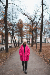 Stylish young woman wear bright pink jacket walking in autumn park outdoors. Fall season.