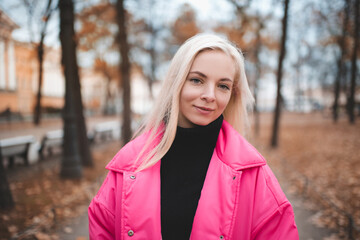 Smiling blonde young woman wear pink bright jacket in park over autumn background outdoors. Looking at camera. Fall season.