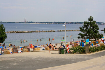View of beach with crowd of people at University of Kiel sailing center in summer with Laboe naval memorial tower and clouds in blue sky background.