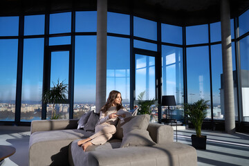 Charming young woman reading magazine while sitting on grey couch with panoramic windows. Bright apartment with modern interior.