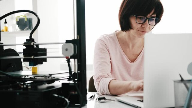 Adult beautiful woman typing on notebook while 3D printer working