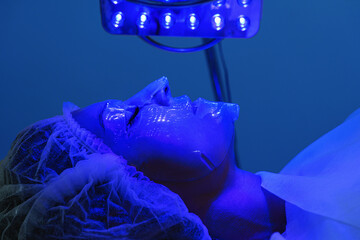 Young woman having blue LED light facial therapy treatment in beauty salon. Beautician wearing face...