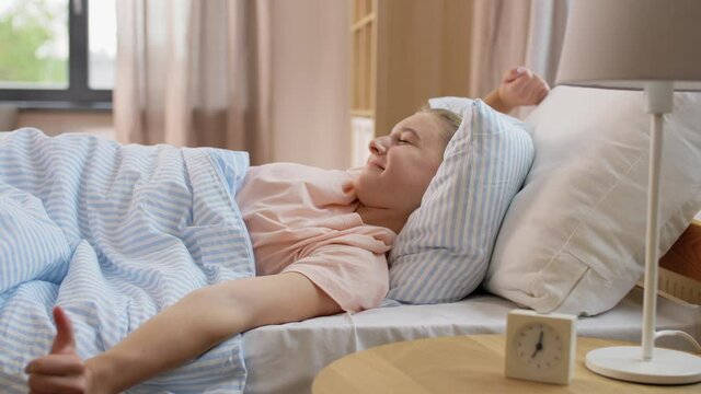 people, bedtime and rest concept - sleepy girl with alarm clock awaking in bed at home