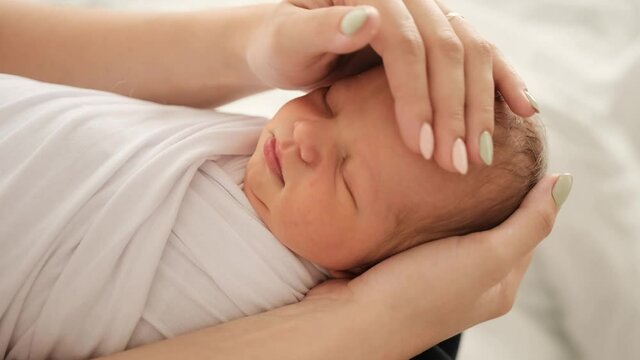 Mother hands strocking head of newborn baby swaddled in white fabric. Girl calming sleeping infant. Little child napping and his mom caring about kid