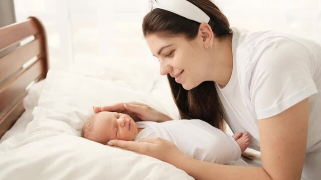 Beautiful young mother strocking head of newborn baby swaddled in white fabric and smiling. Pretty girl calming sleeping infant on the bed. Little child napping and his mom caring about kid