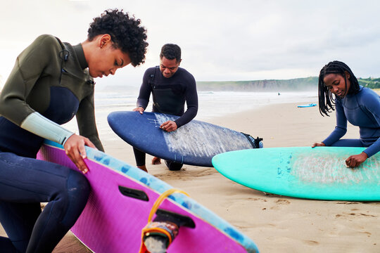 Black Surfers Waxing Colorful Surfboards On Sandy Beach Against Ocean