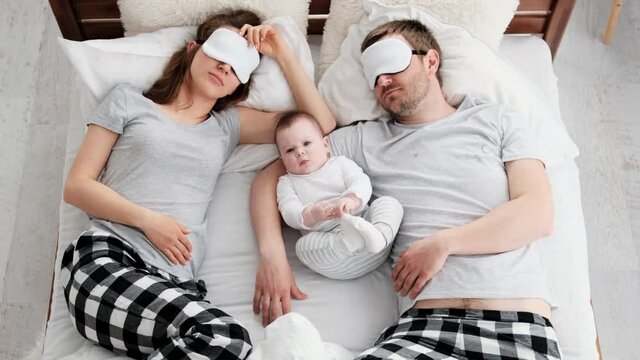Beautiful family wearing same pajamas lying in the bed with little child boy and sleeping. Toddler baby looking at the camera while his young parents sleeping wearing eye masks