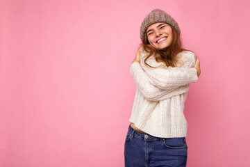 Photo of beautiful happy smiling young brunette woman isolated over pink background wall wearing knitted white sweater and grey warm hat and having fun