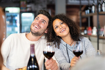 Happy multiracial couple of lover drinking red wine at fashion restaurant - Funny situation with both man and woman looking up - Relationship concept with boyfriend and girlfriend at first date