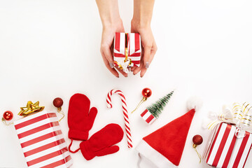 Female hands hold a box with a gift on Christmas flat lay. Christmas decor: gift boxes, candy cane, tree, balls, red mittens and a cap Santa Claus. Red and white classic Christmas still life.