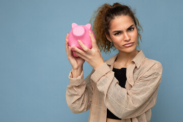 Portrait photo of sincere emotional doubtful cute young beautiful attractive female person with dark curly hair in a ponytail wearing casual beige shirt isolated over blue background with copy space