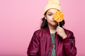 stylish preteen girl in winter outfit holding lollipop and covering eye isolated on pink.