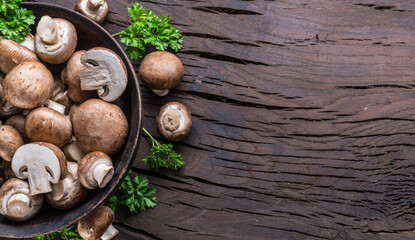Brown colored common mushrooms in wooden bowl on wooden table with herbs. Top view.