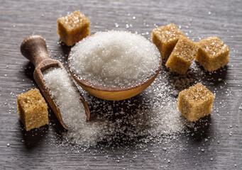 Granulated table sugar in wooden bowl and in the spoon and sugar cubes around it on the table.