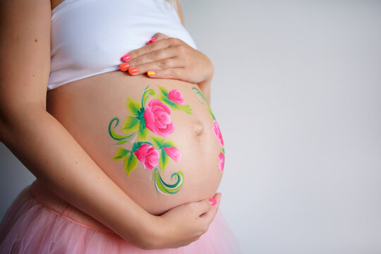 A close-up of the abdomen of a pregnant woman on a light background. On a pregnant belly painted aqua makeup pink flowers with green leaves