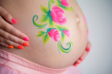 Close-up of a pregnant belly, on which the artist paints with a brush aqua makeup of pink flowers with green leaves. High quality photo