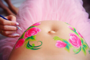 Obraz na płótnie Canvas Close-up of a pregnant belly, on which the artist paints with a brush aqua makeup of pink flowers with green leaves. High quality photo
