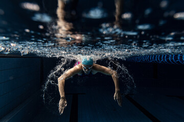 Underwater view of swimming movements details. One female swimmer in swimming cap and goggles training at pool, indoors.