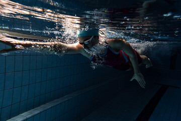 One female swimmer in swimming cap and goggles training at pool, indoors. Underwater view of swimming movements details.