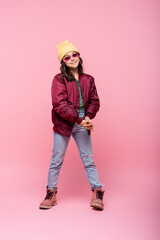 full length of happy preteen girl in trendy outfit and sunglasses on pink.