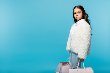 preteen girl in white faux fur jacket holding shopping bags on blue.