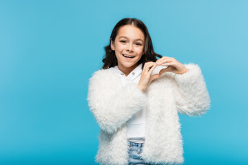 positive preteen girl in white faux fur jacket showing heart with hands isolated on blue.