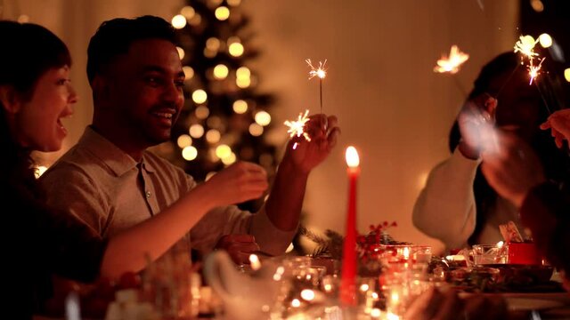 holidays and celebration concept - multiethnic group of happy friends with sparklers having christmas dinner at home over snow