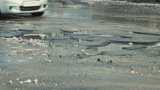 Close-up of cars driving along a broken road with pits and puddles. Large potholes after rain, damaged road infrastructure. Asphalt in violation of technology. Taxpayer money is wasted. UHD 4K.