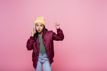 preteen girl in stylish outfit talking on cellphone and pointing with finger isolated on pink.