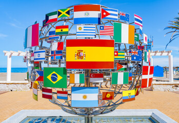 Bandera de España junto a la de muchos países del mundo sobre una esfera de metal simbolizando el planeta tierra y la amistad de España con las demás naciones. - obrazy, fototapety, plakaty