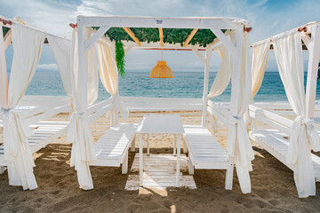 Fototapeta na wymiar Pérgola de madera con cortinas, una mesa y asientos todo de color blanco, con una lampara colgando de mimbre, situada sobre la arena de la playa frente al mar.