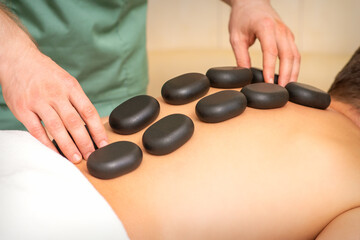 Fototapeta na wymiar Young caucasian man receiving back massage with black stones by masseur in spa salon. A man getting a spa treatment