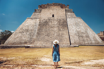 A young woman stands against the backdrop of the Mayan pyramid in Uxmal and enjoys the breathtaking view, from the back. Pyramid of in the ruins of Uxmal, Yucatan, Mexico,Unesco archaeological site - obrazy, fototapety, plakaty