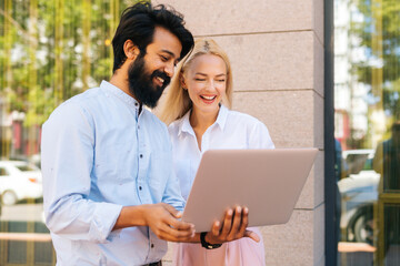 Side view of cheerful bearded business man and young attractive blonde female colleague working on laptop and discuss something outdoors. Businesswoman and businessman talking over laptop.