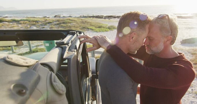 Happy caucasian gay male couple standing by car embracing on sunny day at the beach