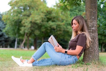 Portrait of beautiful pretty girl reader, young woman, student is reading interesting book in a summer park, sitting on ground, green grass, leaning on tree.