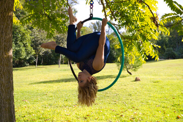 Blonde woman and young gymnast acrobat athlete performing aerial exercise on air ring outdoors in park. Lithe woman in blue costume performs poses of circus performers dancing with hips