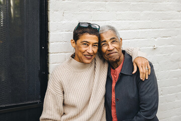 Adult woman and her elderly mother posing together for a portrait against the wall in their backyard