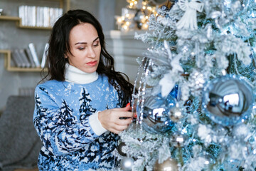 Beautiful brunette girl decorates an artificial christmas tree in blue winter sweater. Merry christmas, new year and winter concept.
