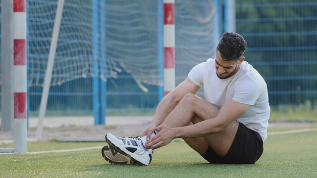 Pain-suffering Middle Eastern footballer sitting on grass of soccer field against goal, holding on ankle, Millennial athlete in summer sportswear Rubs bruised area, upset about injury, cannot continue