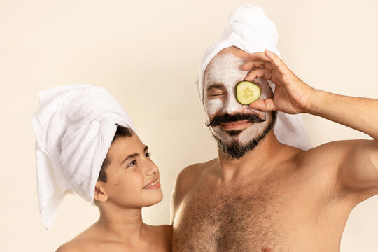 Father And Son On A Facial Spa Day. Caucasian Man With Towel On Head And Cucumber Slice. Boy Looks At His Dad