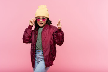happy preteen girl in trendy outfit adjusting sunglasses and pointing at camera isolated on pink.