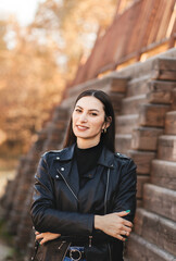 Fototapeta premium A young brunette woman in a black leather jacket in the park in the fall. Walking to the city street.