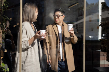 Fototapeta na wymiar Asian businessman and caucasian businesswoman going, talking and drinking coffee inside unknown building. Concept of remote and freelance work. Idea of teamwork and business cooperation. Sunny day