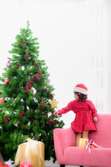 A cute girl in a red polka dot dress and wearing Santa Claus hat stand on the sofa and behind it there is a Christmas tree with twinkling lights and gift boxes under the Christmas tree.