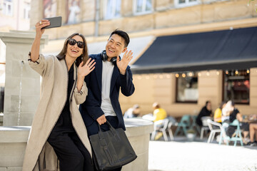 Smiling couple taking selfie on smartphone on city area with fountain. Concept of city life. Idea of friendship. Young beautiful woman and asian man waving hands. Warm sunny day